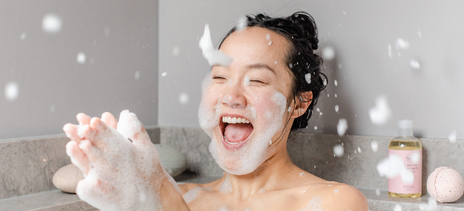Woman laughing and clapping her hand throwing bubbles everywhere in the bath using SLS-free all-natural bubble bath from Rocky Mountain Soap Company