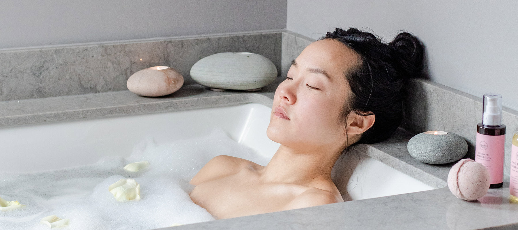 Woman lying in the bath with her eyes closed. There are all-natural sulfate free bubbles from Rocky Mountain Soap Company, a bath bomb and massage oil on the side of the tub. 