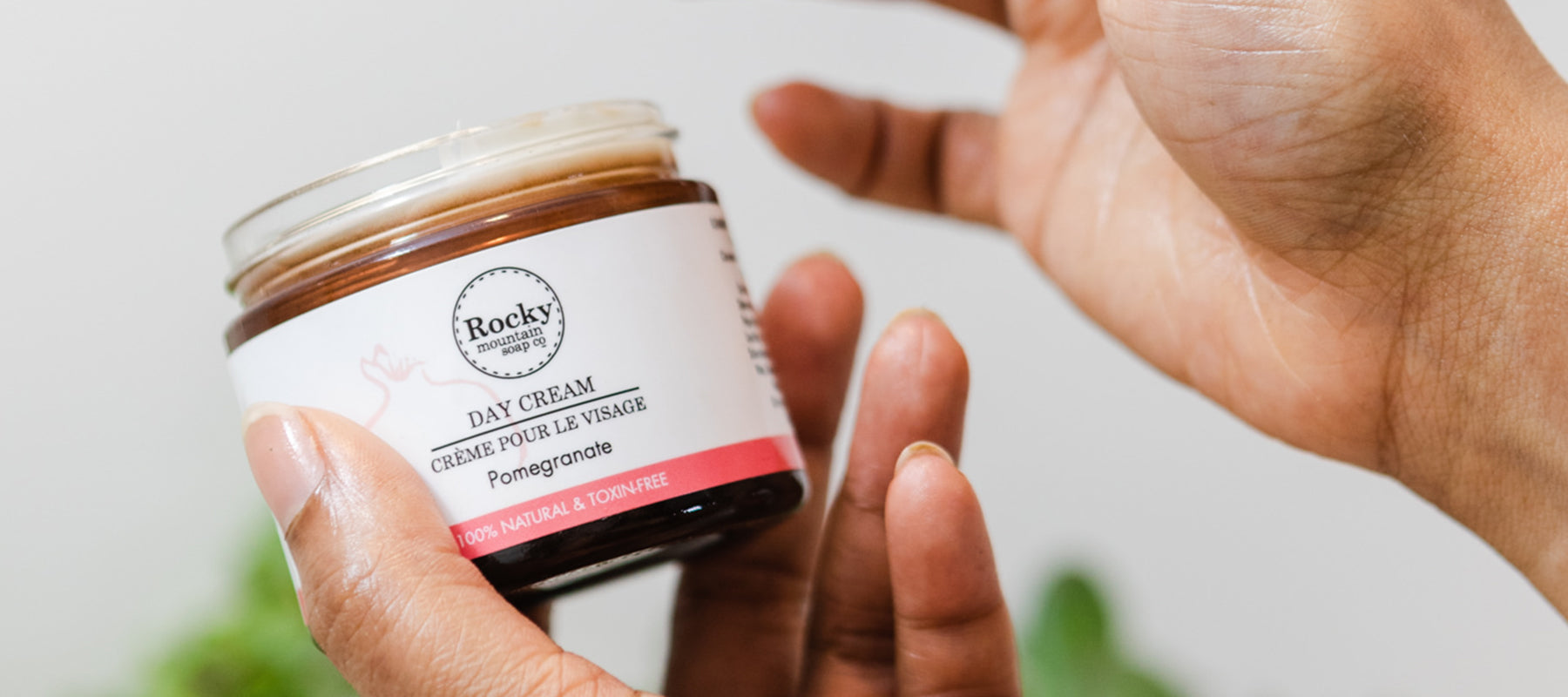 Hand holding an open jar of Rocky Mountain Soap Company Day Cream, a hydrating pomegranate face cream.  
