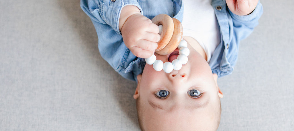 A baby lying on a grey background with a teething ring in their mouth.