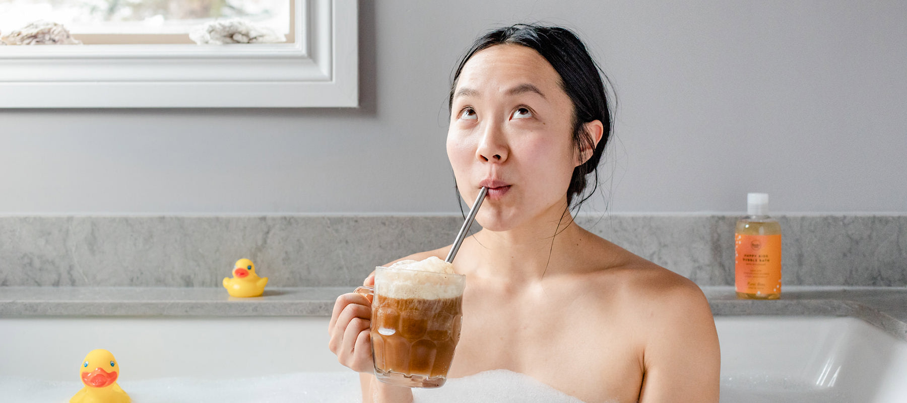 Woman in the bath drinking a root beer float with a bottle of all natural sls-free root beer scented bubble bath and rubber duckies in the background. 