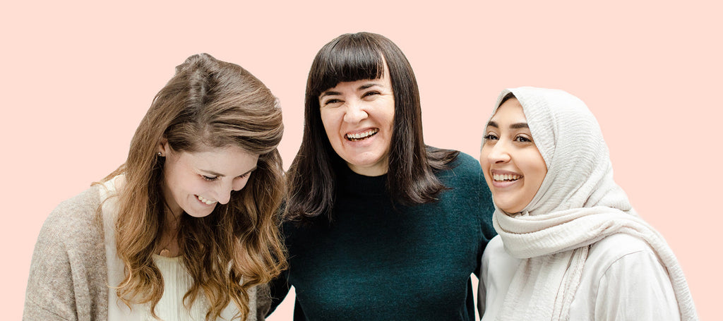 Three women, one with long light brown hair, one with dark hair and bangs, and one wearing a hijab, stand together laughing on a peach background.