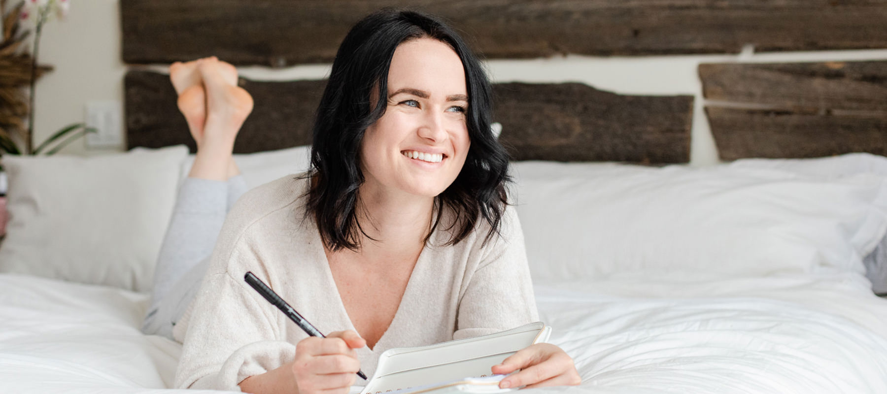 Woman lying on white bed spread writing in her journal. 