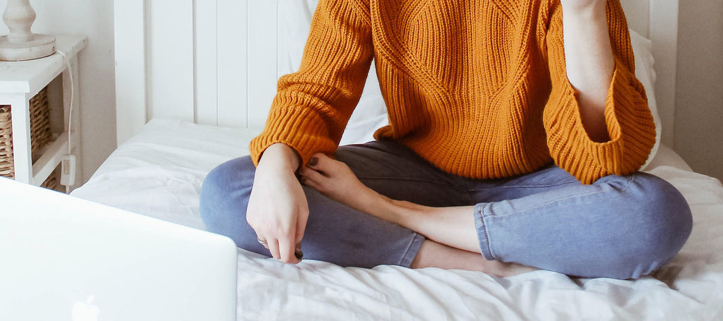 Header image of a woman sitting cross legged on her bed in front of a laptop Rocky Mountain Soap Company how to host a virtual gathering or party during the pandemic blog.