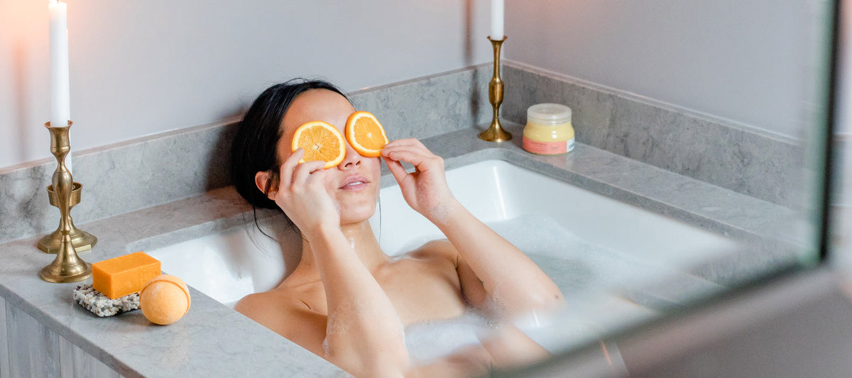 Woman in bath with orange slices on her eyes. On the side of the tub are Canadian local Rocky Mountain Soap Company natural bar soap, bath bomb and body scrub.