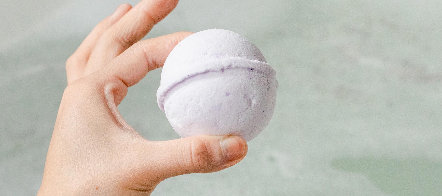 A person's hand holds a white bath bomb between thumb and forefinger, over top of a bath full of water.