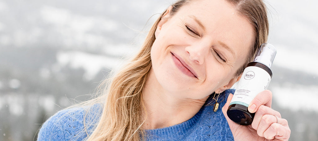 Image of woman smiling holding a bottle of Transformative Cleansing Oil to her face.