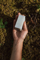A hand holds a rectangular, speckled, pale bar of Minty Tea Tree Soap over a mossy forest floor.