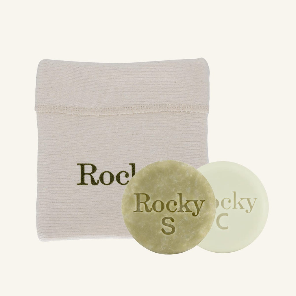 Two round bar soaps, one textured olive green and one smooth pale green, displayed in front of a beige fabric pouch with the brand name 'Rocky' printed on it, against a solid off-white background.