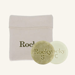 Two round bar soaps, one textured olive green and one smooth pale green, displayed in front of a beige fabric pouch with the brand name 'Rocky' printed on it, against a solid off-white background.