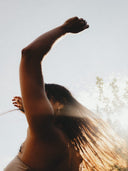 A person with long hair using Scent Free Natural Deodorant from Rocky Mountain Soap Company dancing with their arm raised against a bright sky.