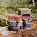 Geranium Soap bars in various colors and patterns are stacked outdoors on a wooden surface with a blurred green background. Some Geranium Soap bars show bubbles, appearing freshly used.