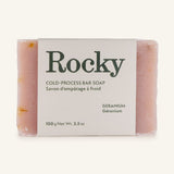 Geranium Soap: a 100g pink handcrafted, cold-process bar with geranium. Partially wrapped in a white label featuring product details in English and French.
