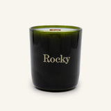 A dark green Moonlight Candle by Rocky Mountain Soap Company in a glass jar with a wooden wick, featuring the word Rocky printed in gold on the front.
