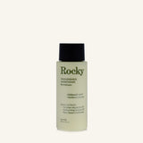 A bottle of Rocky Wild Kindness Conditioner in Rosemary Mint scent, featuring a pale yellow-green translucent bottle, dark green text, and a black cap, set against a solid off-white background.