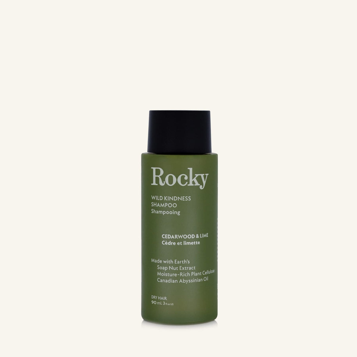 A 90ml bottle of Rocky Wild Kindness Shampoo in Cedarwood & Lime for dry hair, featuring an olive green bottle and a black cap, centered on a light cream background.