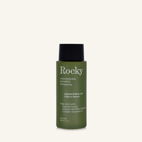 A 90ml bottle of Rocky Wild Kindness Shampoo in Cedarwood & Lime for dry hair, featuring an olive green bottle and a black cap, centered on a light cream background.