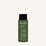 A 90ml bottle of Rocky Wild Kindness Shampoo in Rosemary Mint for normal hair, featuring an olive green bottle and a black cap, centered on a light cream background.