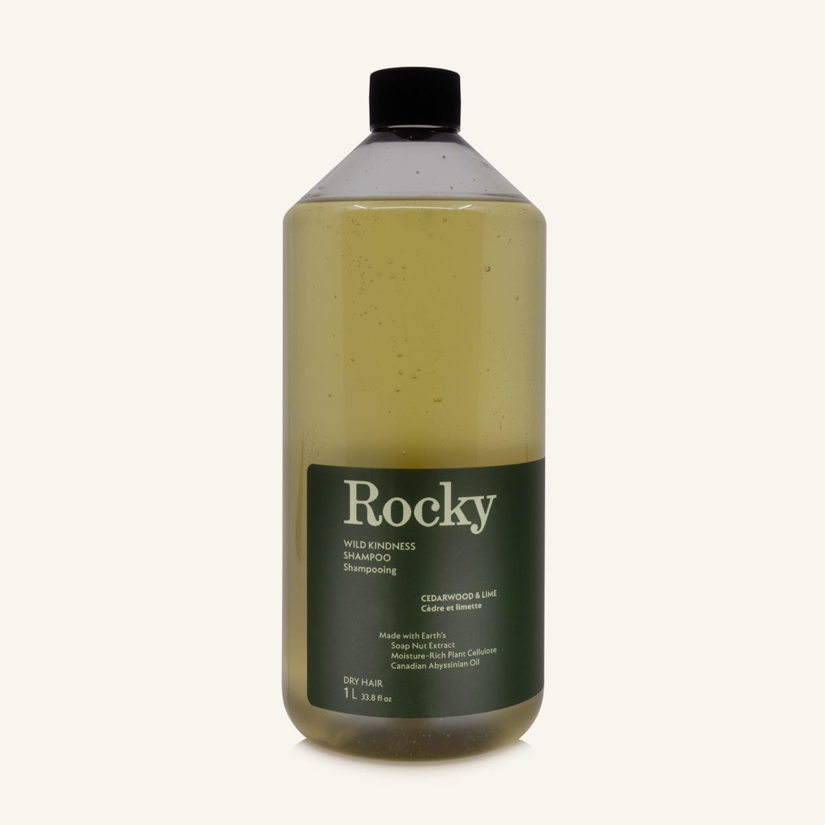 A 1L bottle of Rocky Wild Kindness Shampoo in Cedarwood & Lime for dry hair, featuring an olive green bottle and a black cap, centered on a light cream background.