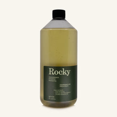 A 1L bottle of Rocky Wild Kindness Shampoo in Cedarwood & Lime for dry hair, featuring an olive green bottle and a black cap, centered on a light cream background.