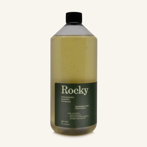 A 1L bottle of Rocky Wild Kindness Shampoo in Cedarwood & Lime for dry hair, featuring an olive green bottle and a black cap, centered on a light cream background.