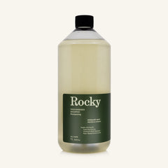 A 1L bottle of Rocky Wild Kindness Shampoo in Rosemary Mint for normal hair, featuring an olive green bottle and a black cap, centered on a light cream background.