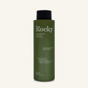 A 360ml bottle of Rocky Wild Kindness Shampoo in Cedarwood & Lime for dry hair, featuring an olive green bottle and a black cap, centered on a light cream background.