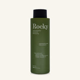 A 360ml bottle of Rocky Wild Kindness Shampoo in Cedarwood & Lime for dry hair, featuring an olive green bottle and a black cap, centered on a light cream background.