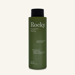 A 360ml bottle of Rocky Wild Kindness Shampoo in Cedarwood & Lime for dry hair, featuring an olive green bottle and a black cap, centered on a light cream background.