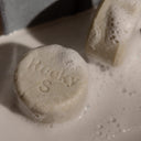 Two round, sudsy Hair Bar: Solid Shampoo bars with strengthening rice protein embossed with text, resting in a white dish.