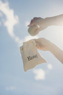 Hands holding a Hair Bar Travel Bundle solid shampoo bar above a small cloth drawstring bag labeled Rocky against a blue sky.