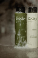 Two bottles of Rocky hair care products—a green Rosemary Mint Wild Kindness Shampoo and a white conditioner—stand side by side with bubbles and water droplets gleaming on their labels.