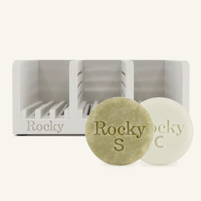 Two round solid hair bars in front of a white divided container for soap, on a light cream background.