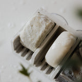A Hair Bar Essentials Bundle shampoo bar and conditioner bar with lather sit in a slatted soap dish.