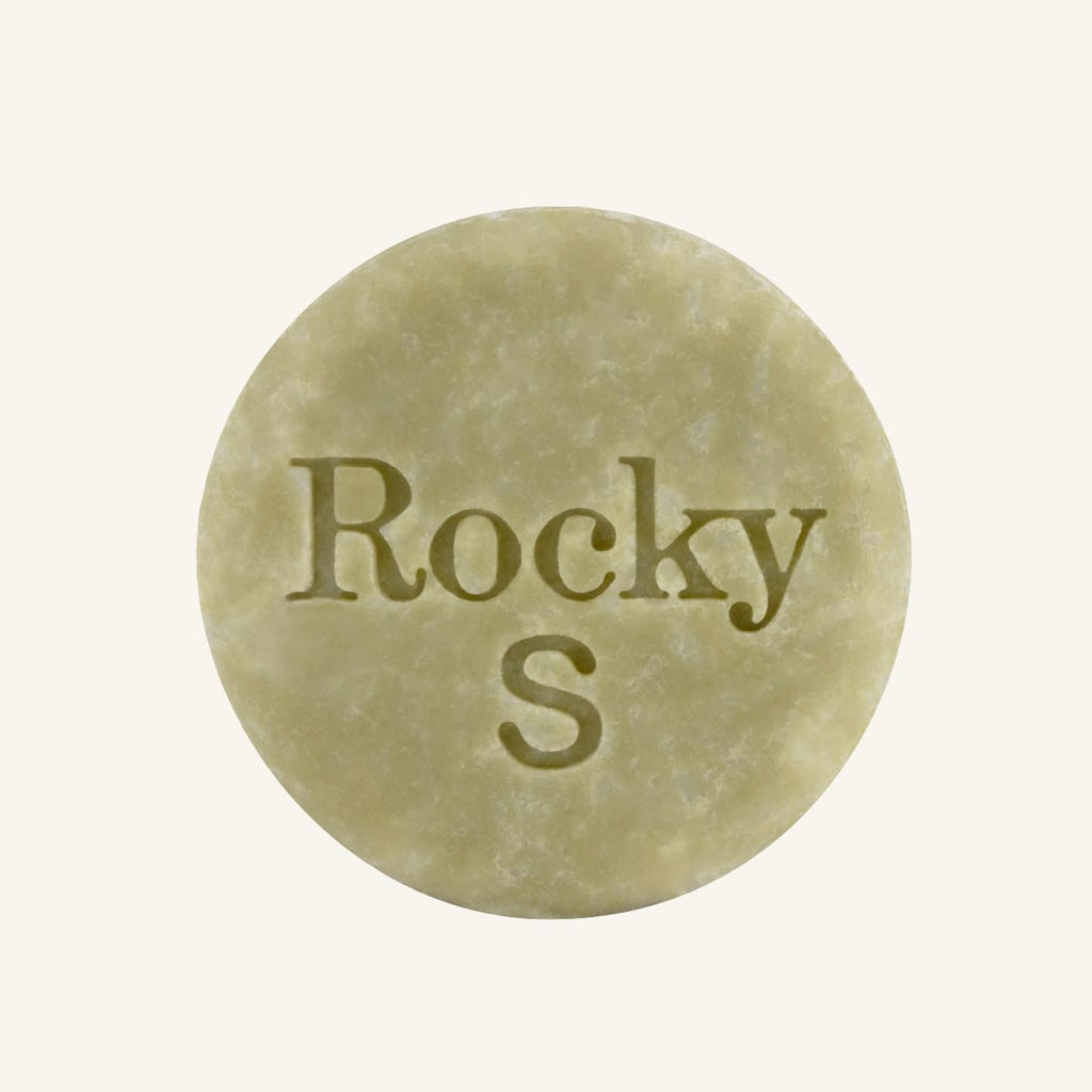 A round, green solid shampoo hair bar embossed with the text 'Rocky S, centered on a solid cream background.
