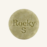 A round, green solid shampoo hair bar embossed with the text 'Rocky S, centered on a solid cream background.