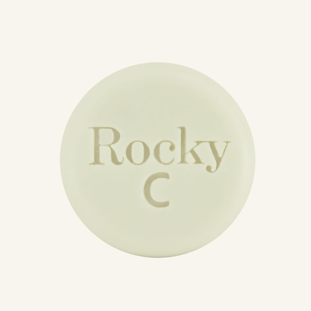 A round, white solid conditioner hair bar embossed with the text 'Rocky C, centered on a solid cream background.