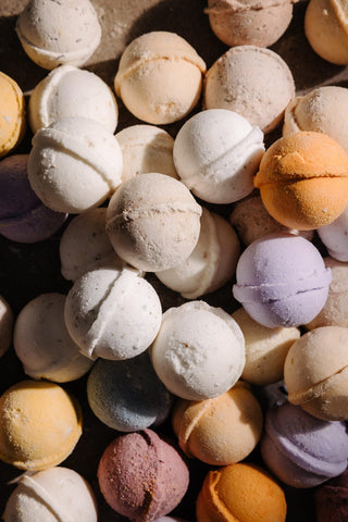 A close-up pile of round bath bombs in assorted pastel and neutral colors, including cream, peach, lavender, pink, and blue-gray, filling the frame.