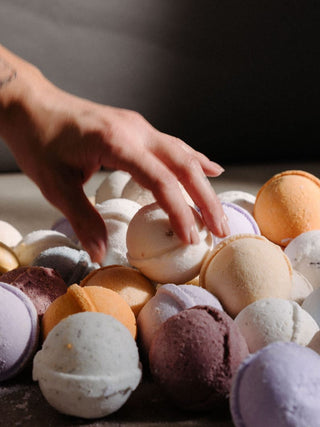 A hand reaches toward a pile of colorful bath bombs. The bath bombs are round and come in pastel shades of lavender, peach, cream, and mauve.